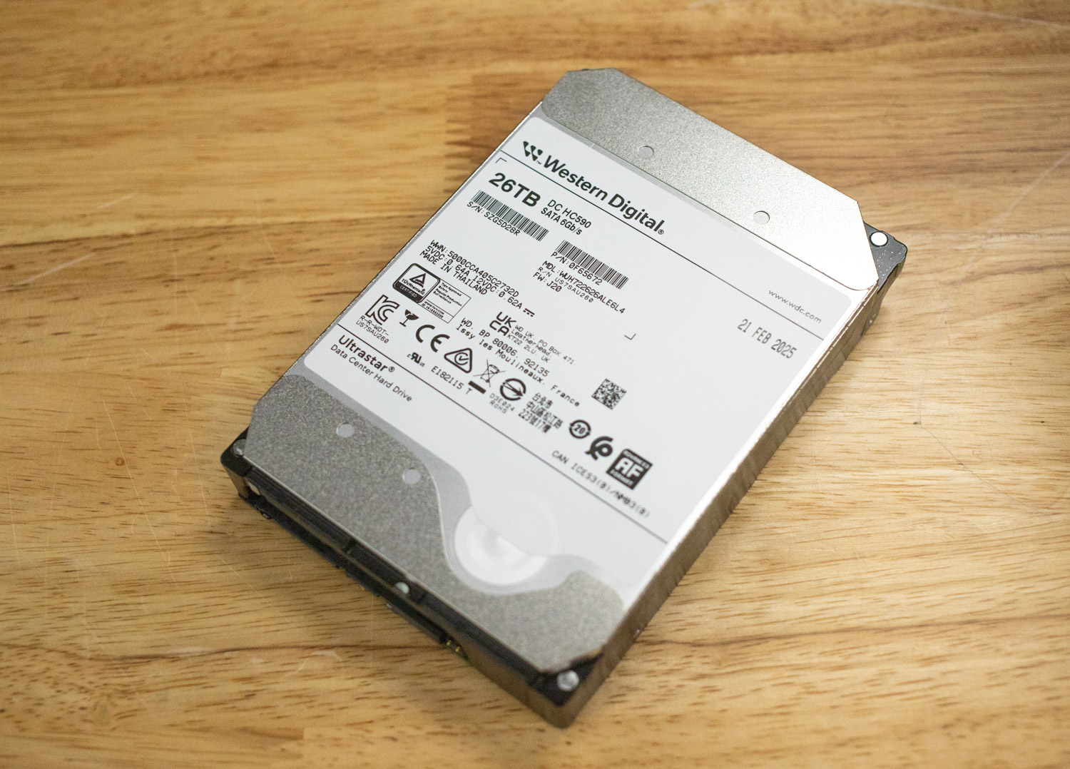 Angled top-down view of a Western Digital Ultrastar DC HC590 26TB hard drive placed on a wooden surface. The drive's white label shows branding, capacity, SATA 6Gb/s interface, model and serial numbers, certifications.